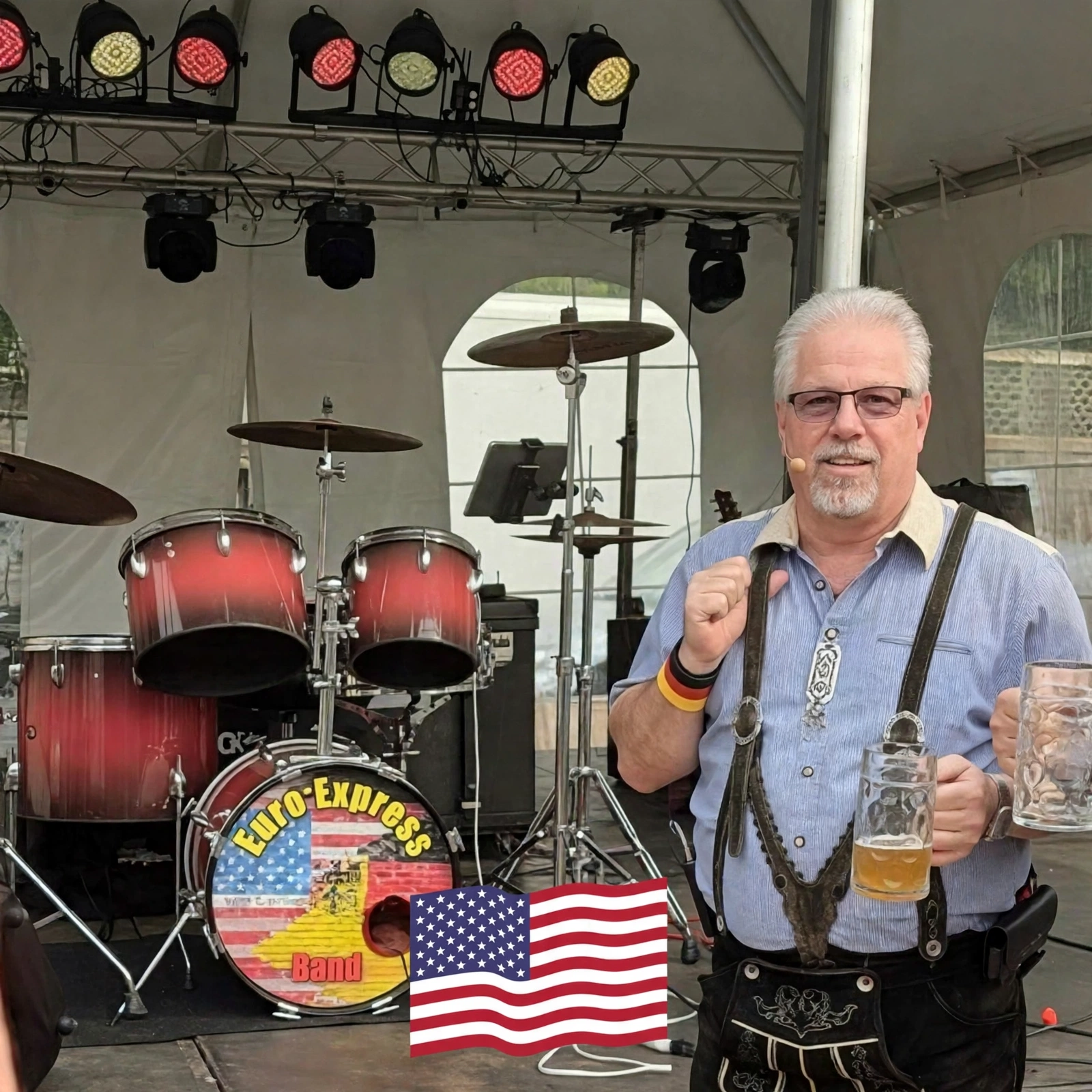 John Gunther of Euro Express Band in lederhosen on an Oktoberfest stage, stein in hand, in front of the band's drum kit