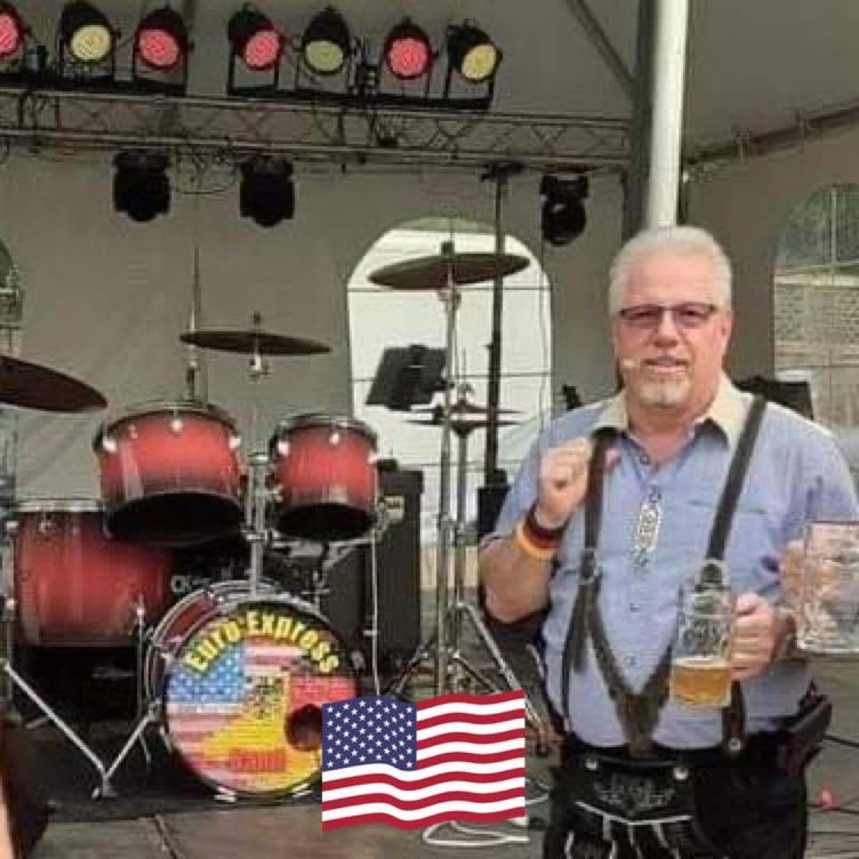 John Gunther of Euro Express Band in lederhosen on an Oktoberfest stage, stein in hand, in front of the band's drum kit